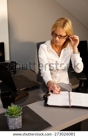 Blonde businesswoman sitting at desk, writing notes to personal organizer, using earbuds.