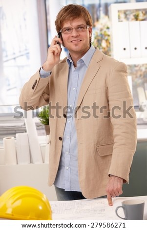 Happy caucasian architect talking on phone at office pointing at table. Wearing glasses, looking up, hardhat.