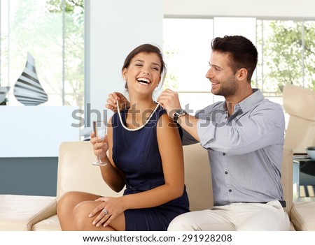 Happy caucasian woman with champagne in hand getting pearl necklace gift from husband. Happy couple, sitting at home on sofa in living room, romance, jewelry.