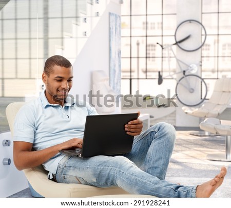 Relaxed casual afro american man browsing online with laptop computer at bright loft apartment. Sitting, barefoot, smiling, looking at screen.