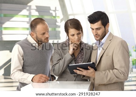 Young caucasian business people with tablet computer at office lobby. Standing, looking at screen, wearing suit.