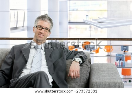 Mature caucasian businessman sitting on sofa at business office. Suit and tie, wearing glasses, smiling, looking at camera.