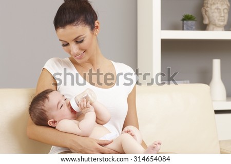 Smiling mother holding naked little baby in arms. Baby drinking from feeding bottle.