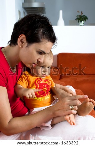 One year old baby and his mother are sitting and resting together on a couch. The mother shows him something. It is a typical home environment.