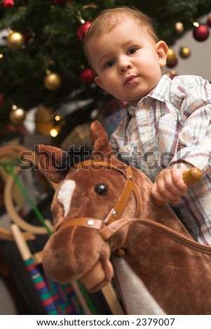 A 1-2 years old child rides his hobby horse under the Christmas tree.
