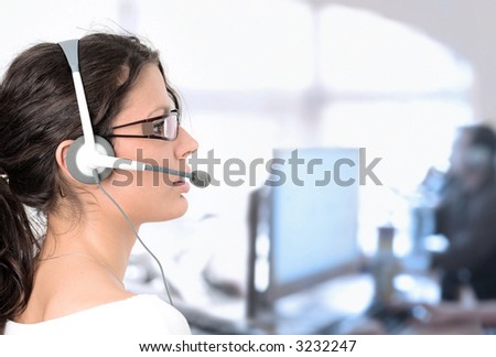 Young female customer service representative recieves calls on a headset while an IT specialist works on a computer in the background.