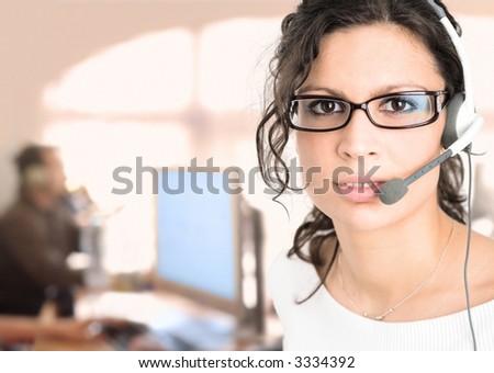 Young female customer service representative receives calls on a headset while an IT specialist works on a computer in the background.