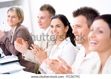 Five business people sitting in a row, smiling and clapping on business training. Selective focus placed on businesswoman in middle.