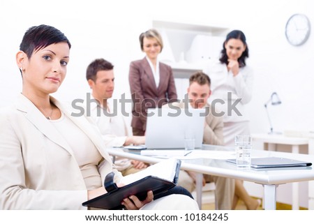 Businesswoman sitting in front, writing notes, looking at camera, smiling. Four business colleagues working on laptop computer in background.