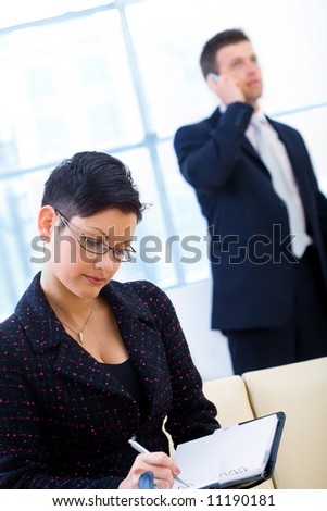 Young and attractive business people working in office. Businesswoman sitting on sofa writing to notebook while businessman calling on mobilephone.