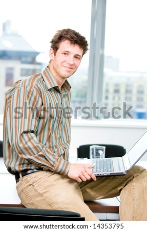 Happy young office worker sitting on table at office and using laptop computer, smiling.