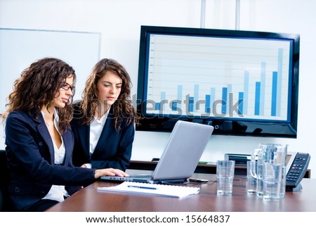 Young businesswomen sitting by meeting table at office in front of a huge blank plasma TV screen and using laptop and phone.