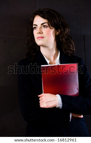 Young businesswoman in dark suit holding red folder daydreaming on office corridor posing for business portrait.