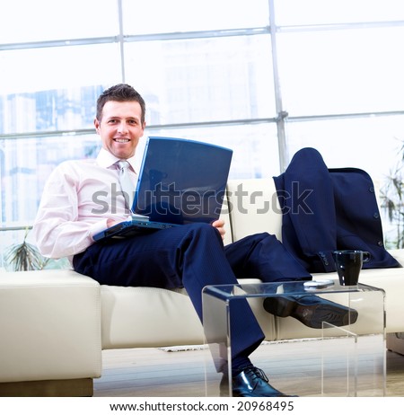 Happy businessman sitting on couch at office, working on laptop computer and smiling.