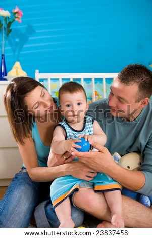 Portrait of happy family at home. Baby boy ( 1 year old ) and young parents father and mother sitting on floor and playing together at children's room, smiling.