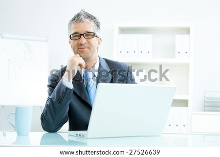 Businessman with grey hair, wearing grey suit and glasses thinking over laptop computer, sitting at office desk leaning on hand, smiling.