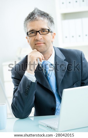 Businessman with grey hair, wearing grey suit and glasses thinking over laptop computer, sitting at office desk leaning on hand.