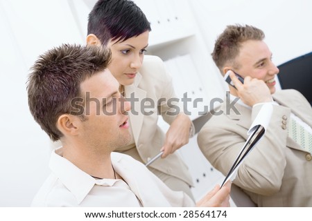 Business team working together in office. Man and woman reviewing notes in the foreground, happy man talking on mobile phone in the background.