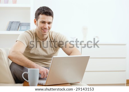 Happy young man in t-shirt sitting on sofa at home, working on laptop computer, smiling.