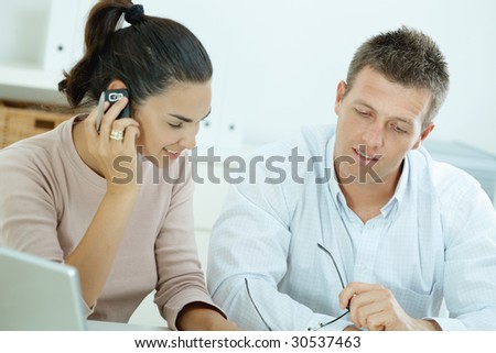 Couple working on laptop computer at home office, happy, smiling. Woman calling on mobile phone.
