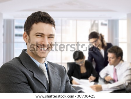 Closeup portrait of happy businessman, looking at camera, smiling. Business team working in the background, sitting at desk.
