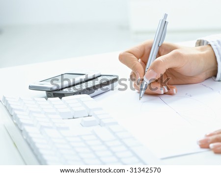 Female hand holding pen, writing on paper, beside desktop computer keyboard and mobile phone.