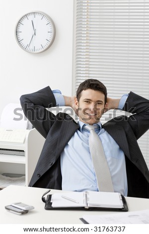Businessman sitting at office desk with satisfied expression, looking at camera, smiling.