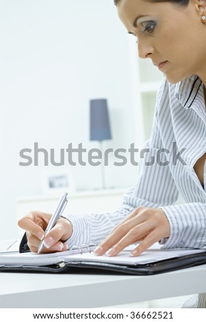 Young woman sitting at desk, writing notes into personal organizer.