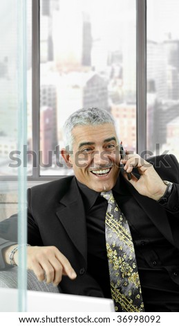 Mature businessman talking on mobile phone in front of office windows, laughing.