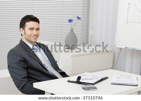 Young businessman sitting relaxed behind office desk with personal organizer, documents and mobil phone.