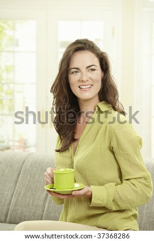 Happy young woman drinking coffee, sitting on couch in living room, smiling.