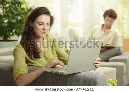 Couple at home. Woman using laptop computer, man reading book in background.
