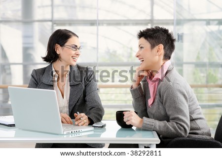 Happy young businesswomen working together at desk in modern office, looking at each other, laughing.