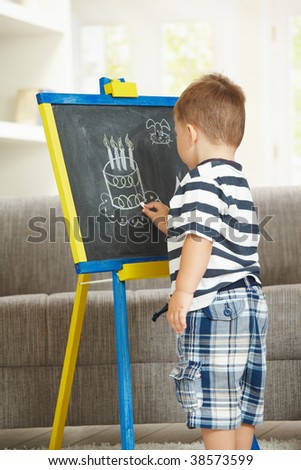 Little boy drawing birthday cake with chalk on blackboard at home.