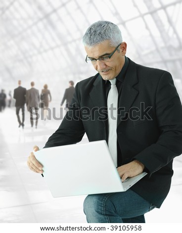Mature businessman  using laptop computer standing in office hallway, holding the machine on his knee.