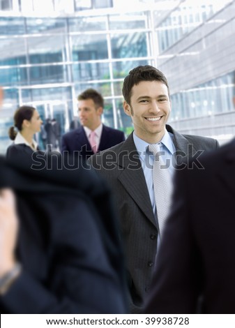 Young businessman walking in crowd in office lobby, smiling.