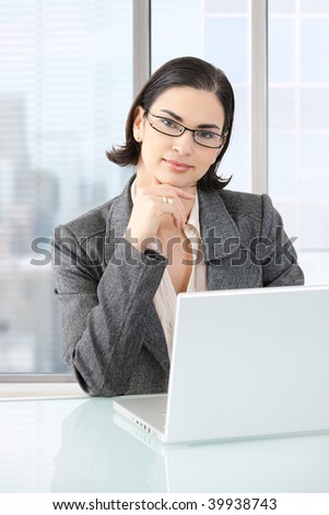 Businesswoman sitting at desk in front of offive windows, thinking over laptop computer.