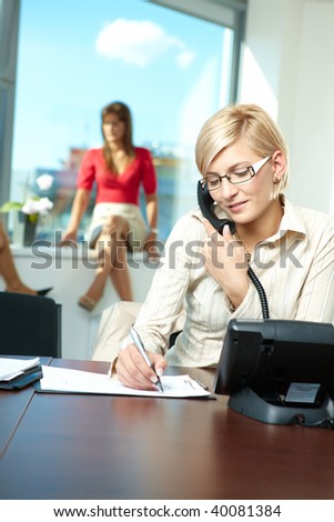 Young businesswoman sitting at desk in office, talking on landline phone, writing notes to personal organizer.