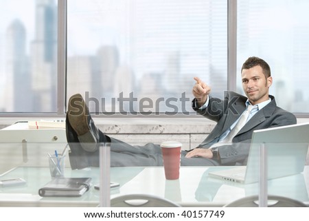 Relaxed businessman sitting at desk in front of office windows, pointing and looking at camera.