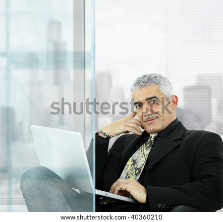 Portrait of mature businessman sitting  in front of windows in office, using laptop computer, smiling.