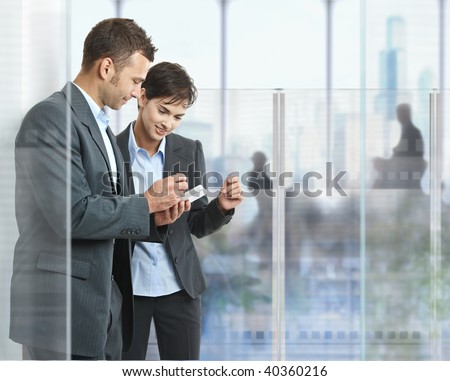 Two businesspeople standing in modern office with glass walls, looking at smart mobile phone.