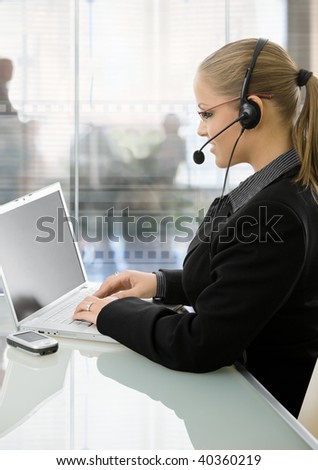 Young businesswoman sitting at desk in modern office with glass walls, using laptop computer, talking on headset.