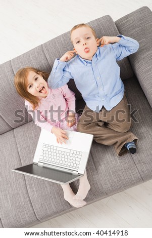 Overhead shot of children sitting on couch at home holding laptop computer, looking up with mocking expression.