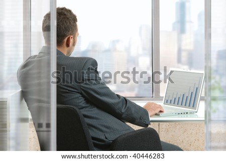 Businessman sitting at desk in office, working with laptop computer, looking out the windows.