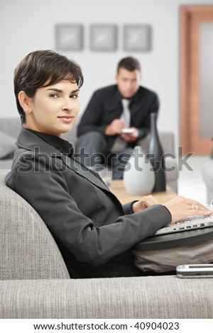 Young attractive businesswoman sitting on sofa at office lobby working with laptop computer, smiling.