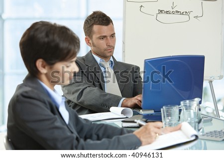 Businessman and businesswoman sitting at desk in office, using laptop computer and writing notes.