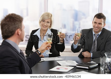 Happy young business people sitting around meeting table at office celebrating success with champagne, smiling.