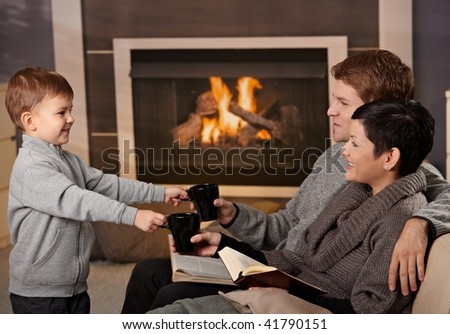 Happy family sitting on couch at home in front of fireplace, drinking tea, smiling.