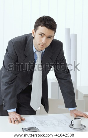 Architect wearing grey suit leaning on office desk with floor plan on it. Looking at camera.