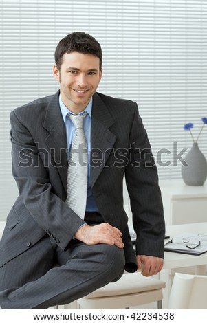 Portrait of businessman wearing grey suit and blue shirt, sitting on office desk, smiling.